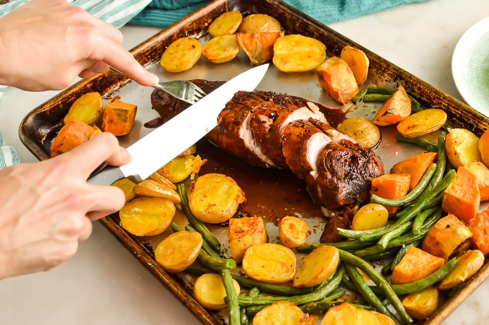 Woman's hands slicing brown sugar balsamic roasted pork on a tray with potatoes, sweet potatoes, and green beans.