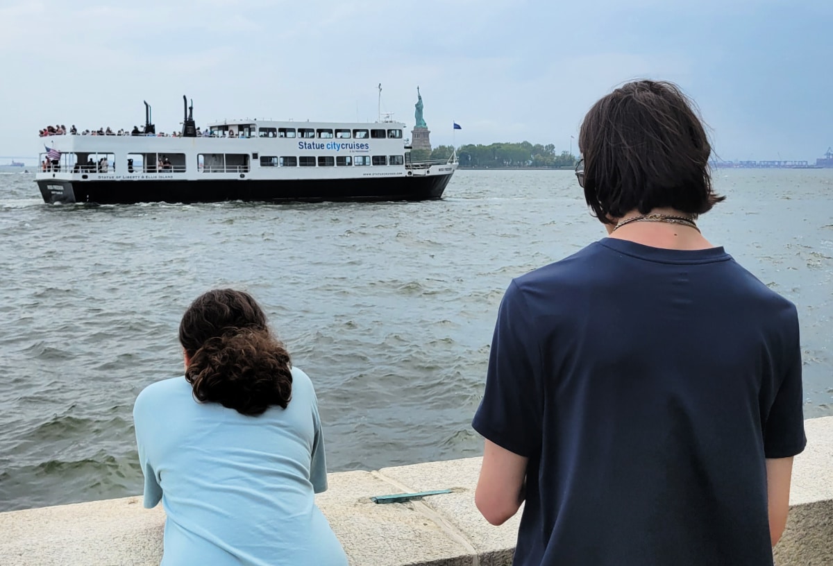 Two teens look at the Ellis Island and Liberty Island ferry as it sails past the Statue of Liberty.