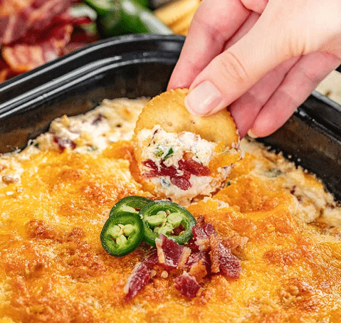 Fingers holding a cracker with jalapeno popper dip over a bowl.
