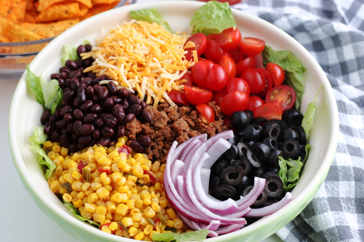 How to Make Dorito Taco Salad: bowl of tomatoes, black beans, tomatoes, cheese, corn, onions, olives, and beef.