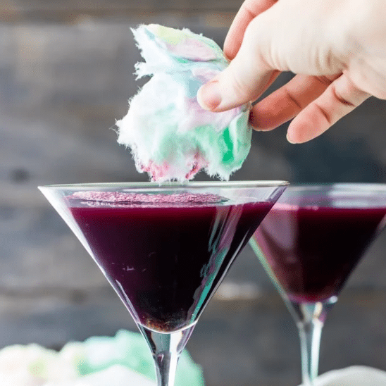 Hand holding cotton candy above a martini glass of full of a grape cocktail.