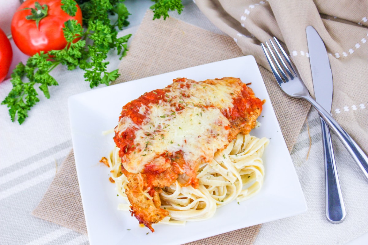 Top down view of Chicken Parmesan Fettuccine Alfredo on a plate next to a fork and knife.