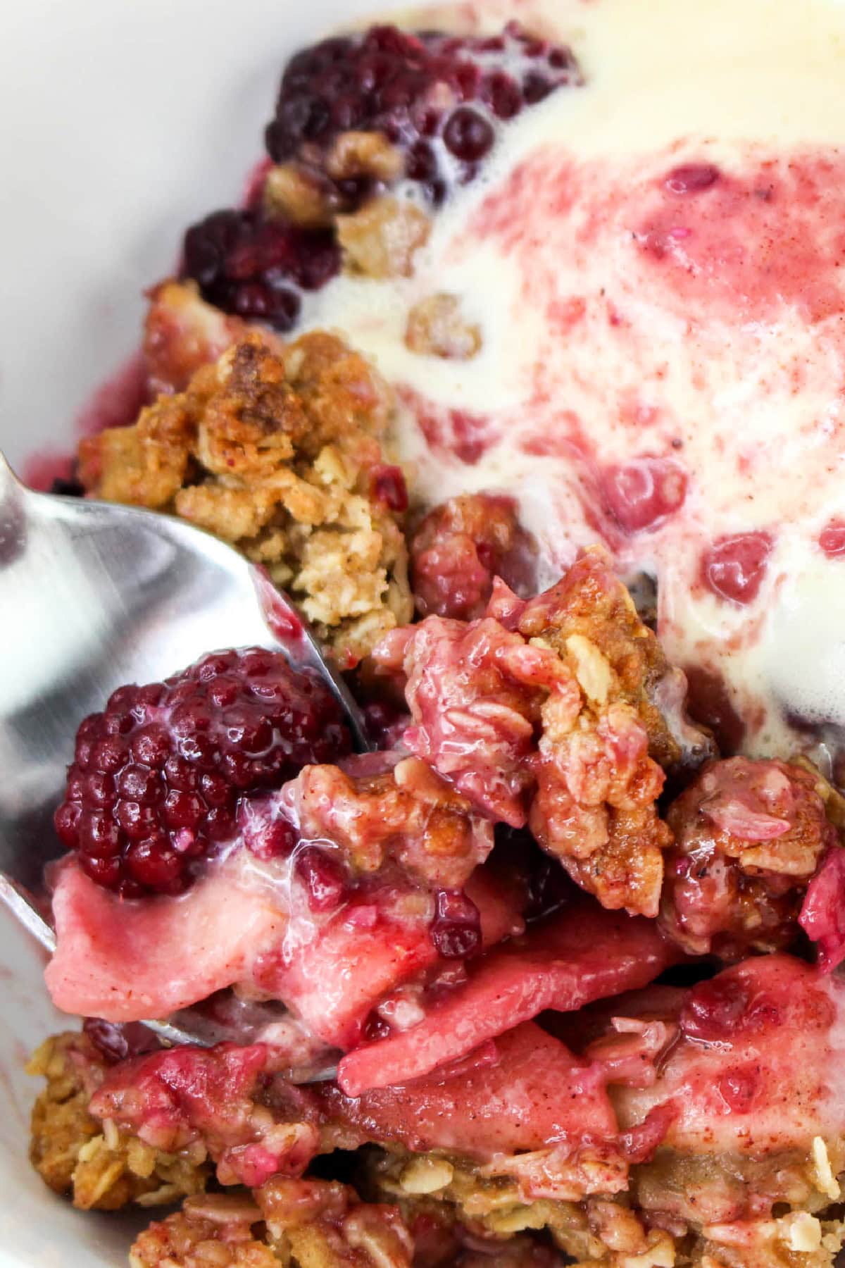 Close up of Apple and Blackberry Crumble with metled ice cream and a spoon in a bowl.