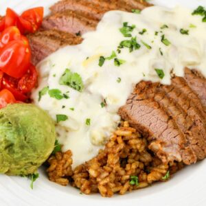 Close up view of a rice bowl with steak, guacamole, chopped tomatoes, and queso sauce.